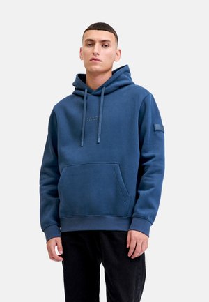 Young man wearing a blue hooded sweatshirt with front pocket and black pants, standing against a plain light gray background.