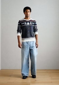 Muž stojaci v svetlomodrých džínsach s širokými nohavicami, čiernych topánkach a tmavom vzorovanom svetri s bielymi motívmi sobov a vločiek.