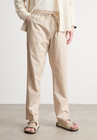 Beige, lette bukser med elastisk liv og justerbar snor. Har sidelommer og en avslappet passform. Brukt med enkle sandaler.