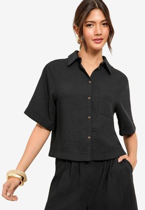 Camicia nera a maniche corte con tessuto testurizzato, chiusura con bottoni, colletto e taschino sul petto. Indossata con pantaloni neri abbinati.