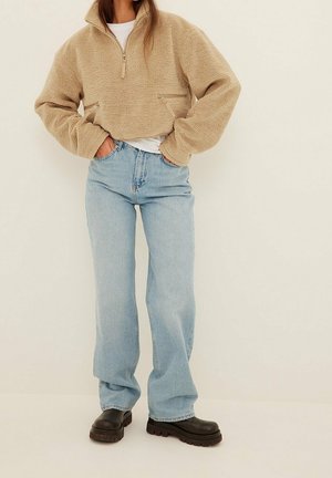 Kvinde iført beige fleece-trøje med lynlås, lyseblå højtaljede vidde-benet jeans og sorte chunky støvler, stående med hænderne i lommerne.