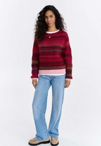 Sweter w paski z dzianiny w kolorach czerwonym i różowym, z długimi rękawami i ściągaczem u dołu, zestawiony z jasnoniebieskimi rozszerzanymi dżinsami i beżowymi butami.