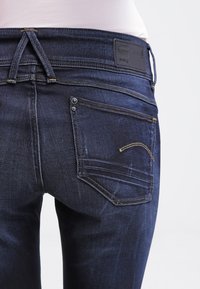 Jeans de ganga azul escura com um design justo, apresentando uma cintura reta, bolsos traseiros costurados e costura em destaque dourada.