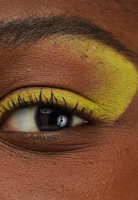Nærbilde av et øye med livlig gul øyenskygge på øyelokket, som står i kontrast til mørkebrun hud og naturlige vipper. Glatt tekstur synlig.