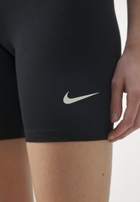 Pantalones cortos atléticos ajustados negros con el logo blanco de Nike en forma de swoosh en el muslo, mostrados en la pierna y mano de una persona.