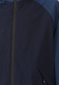 Marinejakke med en glat stofoverflade, frontlynlås og en kontrasterende blå overdel med diskret logodetaljering.