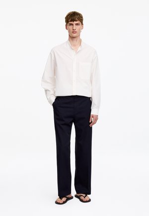 Homme portant une chemise blanche ample boutonnée, un pantalon noir et des tongs noires, debout devant un fond blanc.