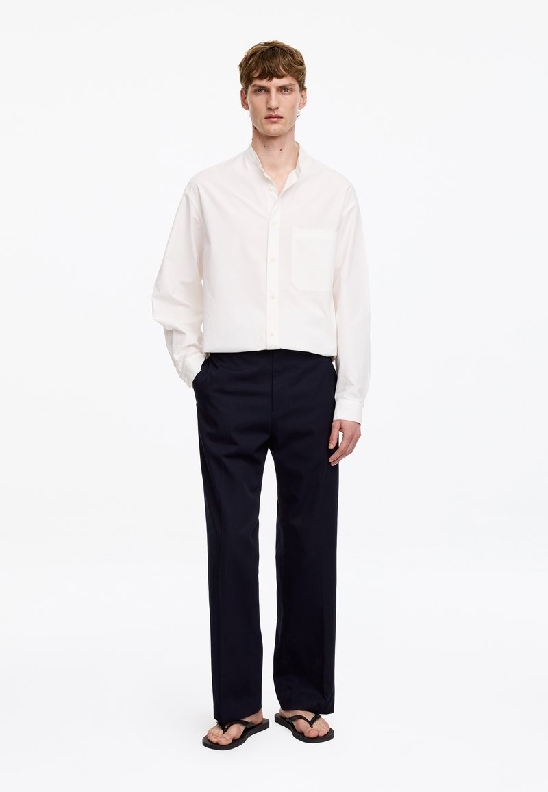 Homme portant une chemise blanche ample boutonnée, un pantalon noir et des tongs noires, debout devant un fond blanc.