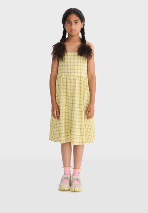 Niña de pie con un vestido sin mangas amarillo de cuadros, calcetines rosas con volantes y zapatillas beige sobre un fondo claro y liso.