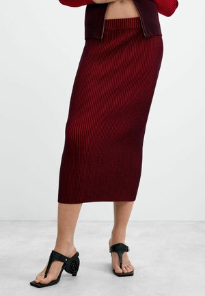 Donna che indossa una gonna midi a righe verticali rosse e nere con sandali neri con tacco e infradito, in piedi su un pavimento grigio chiaro.