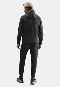 Tuta sportiva grigio scuro con cappuccio e pantaloni jogger coordinati. Tessuto strutturato, vestibilità sfiancata e cuciture discrete. Scarpe sportive nere e rosse.