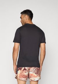 Mann sett fra bak med en ensfarget svart t-skjorte med korte ermer og rutete, flerfargede shorts mot en hvit bakgrunn.