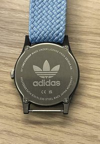 Rokassprādze ar zilu austu siksniņu un nerūsējošā tērauda aizmuguri, kurai ir iegravēta Adidas zīme un citas tehniskās specifikācijas.