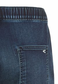 Mørkeblå denim jeans har en rynket talje og en baglomme med et lille logo. Teksturen er glat med en let glans.