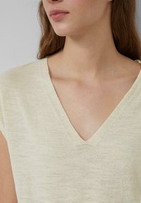 Jasno beżowy sweter z dekoltem w serek, z krótkimi rękawami, miękką teksturą i luźnym krojem. Materiał wydaje się lekki i przewiewny.