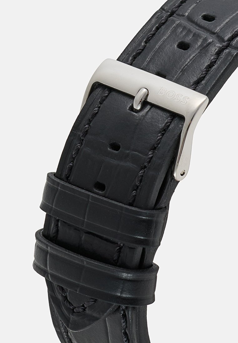Zwarte leren horlogeband met een krokodillenpatroon, zilveren metalen gesp met de merknaam. Meerdere verstelbare gaten zichtbaar.
