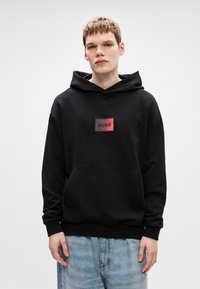 Μαύρο φούτερ με κουκούλα, διαθέτει τσέπη μπροστά και ένα ορθογώνιο λογότυπο με την ένδειξη "HUGO" σε κόκκινο και μαύρο χρώμα. Φτιαγμένο από μαλακό υλικό με άνετη εφαρμογή.