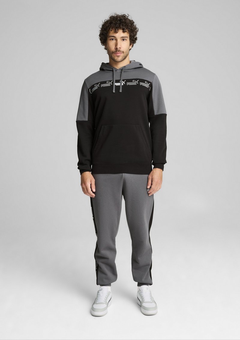 Schwarzer und grauer Trainingsanzug mit einem Kapuzensweatshirt, das eine Kängurutasche und ein farblich unterteiltes Design aufweist. Passende Hose mit seitlichen Akzenten.