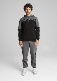 Felpa con cappuccio nera e grigia con tasca frontale e dettaglio logo, abbinata a pantaloni della tuta grigi con strisce laterali, indossata con scarpe da ginnastica bianche.