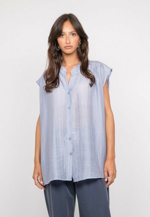 Femme aux longs cheveux foncés portant un chemisier bleu clair sans manches à boutons et un pantalon large foncé, debout devant un fond blanc uni.