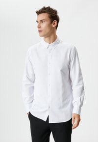 Camisa blanca de manga larga con un sutil patrón de puntos, cuello abotonado y corte regular, presentada con una mano en un bolsillo.