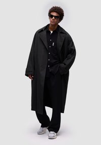 Abrigo largo negro con un corte relajado sobre una camisa negra, combinado con unos pantalones negros holgados y zapatillas grises. Presenta gafas de sol y un diseño minimalista.