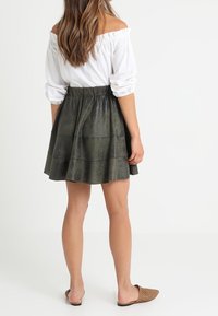Gonna plissettata verde realizzata in un materiale testurizzato, con una vita arricciata. Indossata con una maglietta bianca scollata e scarpe basse.