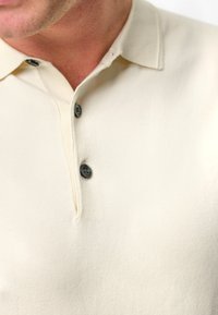 Krämfärgad polo t-shirt i mjukt tyg, med en tvåknappsslutning i svart och en klassisk krage.