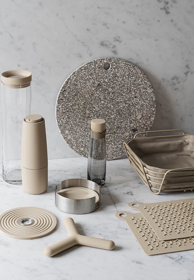 Verschiedene Küchenutensilien, darunter eine Glasflasche, ein beiger Behälter, ein strukturiertes Steinbrett und beige Silikonmatten, auf einer Marmoroberfläche angeordnet.