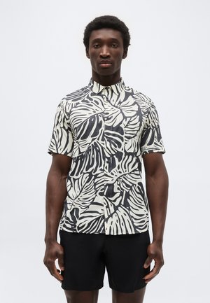 Mann som har på seg en kortermet skjorte med store svarte og hvite bladmønstre og ensfargede svarte shorts mot en ensfarget bakgrunn.