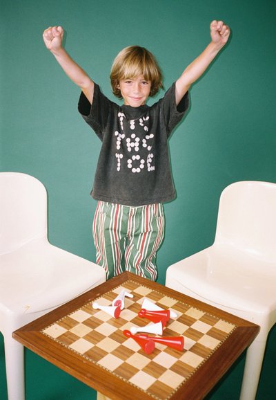 Kind gekleed in een zwart t-shirt met witte tekst, gestreepte groene broek, staat met opgetilde armen naast een houten speltafel en witte stoelen.