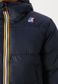 Giubbotto imbottito blu navy con chiusura a zip, strisce multicolori, cappuccio e logo sul petto. Il materiale sembra liscio e lucido.