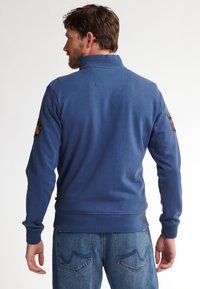 Felpa blu con colletto alto, polsini a costine e dettaglio sul dorso. Presenta toppe ricamate sulle maniche, abbinata a jeans di denim.