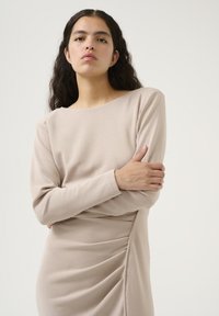 Béžové dlouhé šaty s lodním výstřihem a bočním drapovaným detailem. Měkká, hladká tkanina s přiléhavým siluetou, minimální švy.