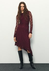Bordeauxrotes Spitzenkleid mit langen Ärmeln und asymmetrischem Saum, kombiniert mit schwarzen kniehohen Stiefeln. Strukturierte Blumenmuster überall.