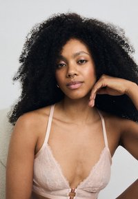 Bralette en dentelle rose clair avec des bretelles ajustables et un fermoir décoratif à l'avant ; caractérisé par des motifs floraux et une texture douce.