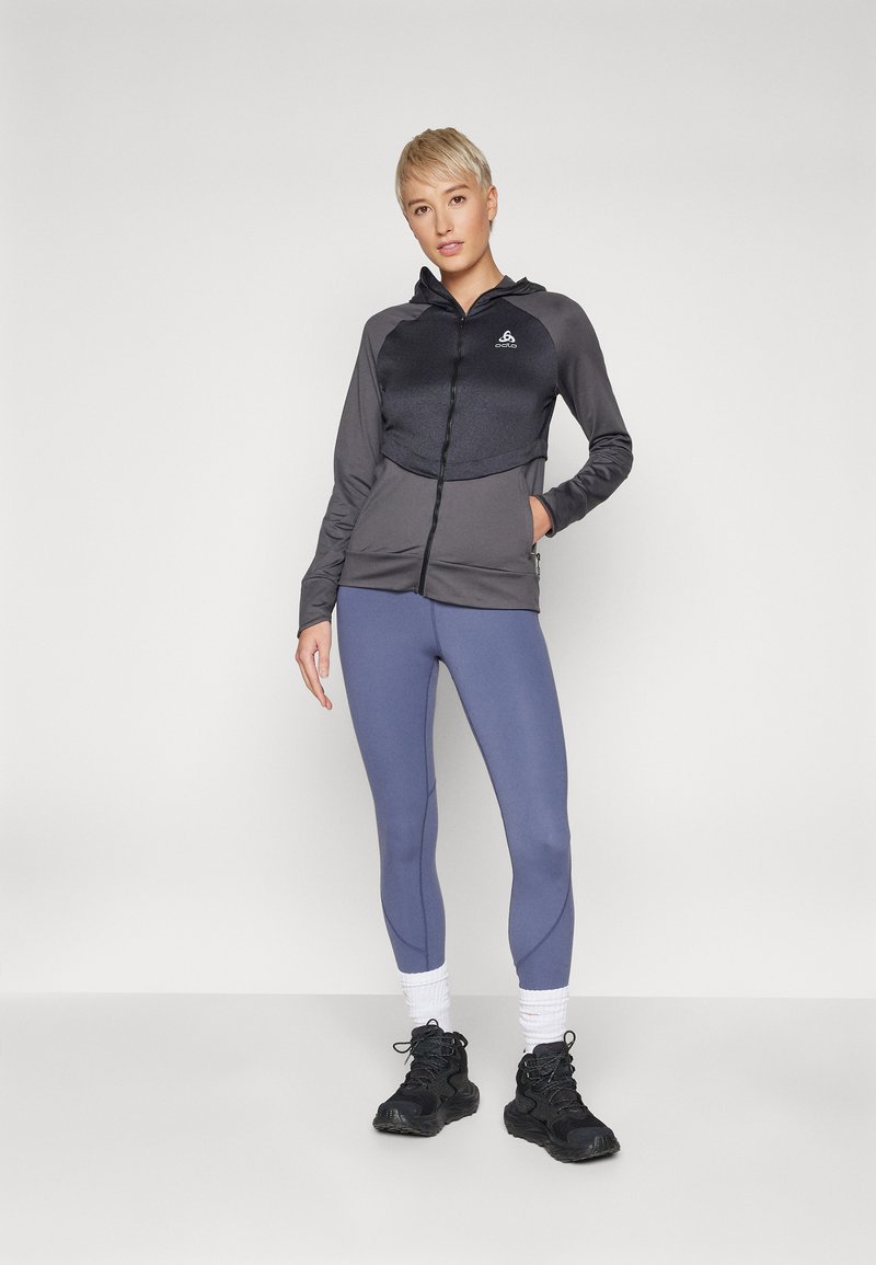 Kapuzensjacke in dunkelgrau mit glänzender Textur, kombiniert mit hellgrauen Leggings und schwarzen Sportschuhen.