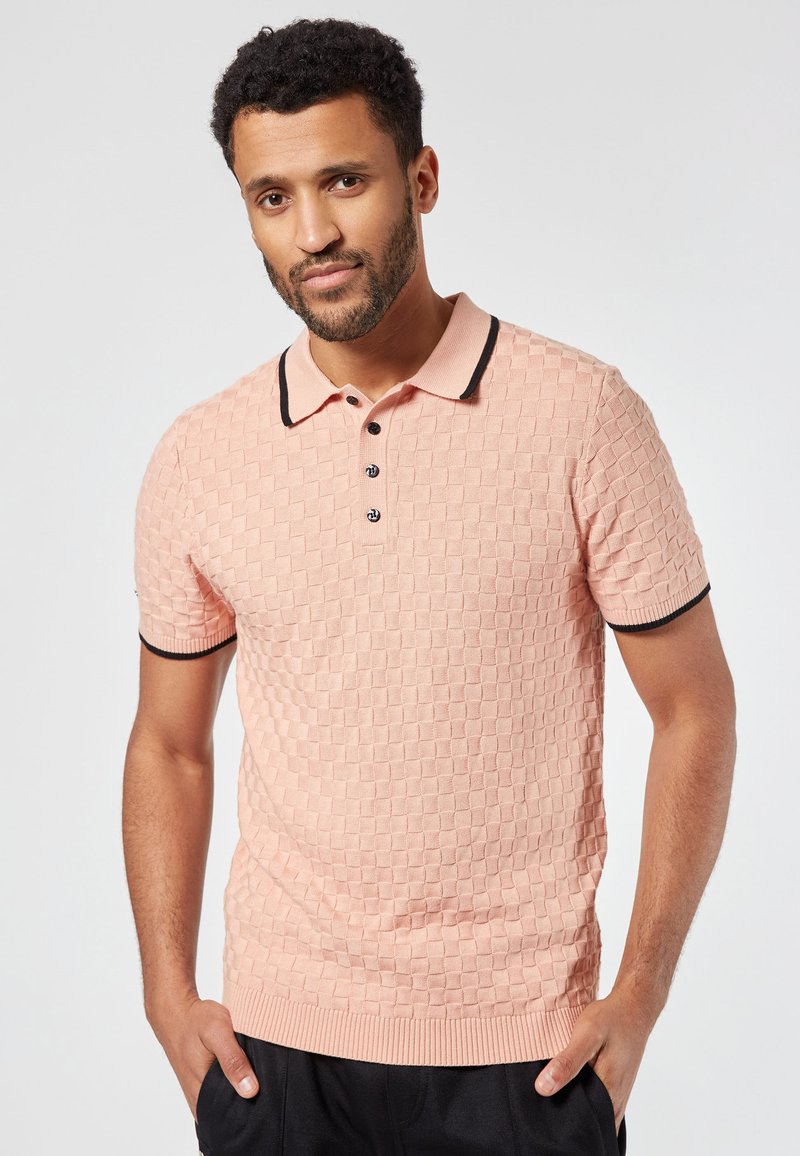 Vaatepaita vaaleanpunaisesta neulotusta kankaasta, jossa on teksturoitu timanttikuosi, musta kaulus ja hihansuut, kolmen napin listakuvio ja tiukka muotoilu.