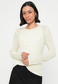 Γυναίκα με σκούρα μαλλιά μέχρι τους ώμους, ντυμένη με μπεζ μπλούζα μακρυμάνικη και μαύρη φούστα, ποζάροντας μπροστά σε λευκό φόντο.