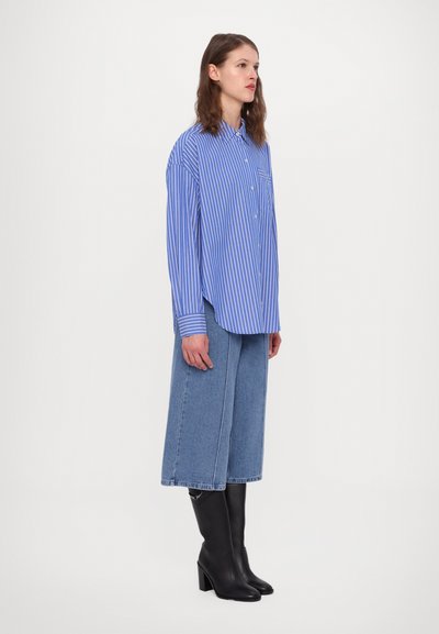 Blue and white striped button-up shirt, relaxed fit, with a chest pocket. Paired with wide-leg denim trousers and tall black boots.