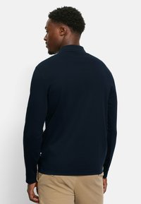 Marineblauwe polo met lange mouwen, gemaakt van zachte stof, met een kraag en een ontspannen pasvorm, en subtiele accenten aan de zijkanten bij de zoom.