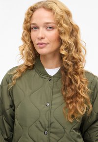 Vadderad olivgrön jacka med rundad krage och tryckknappsförslutning, med slät yta och kurvade sömmönster.