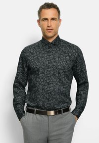 Schwarzes Langarmhemd mit weißem Blumenmuster, ergänzt durch einen klassischen Kragen und einen durchgehenden Knopfverschluss, getragen mit grauen Hosen und einem Gürtel.