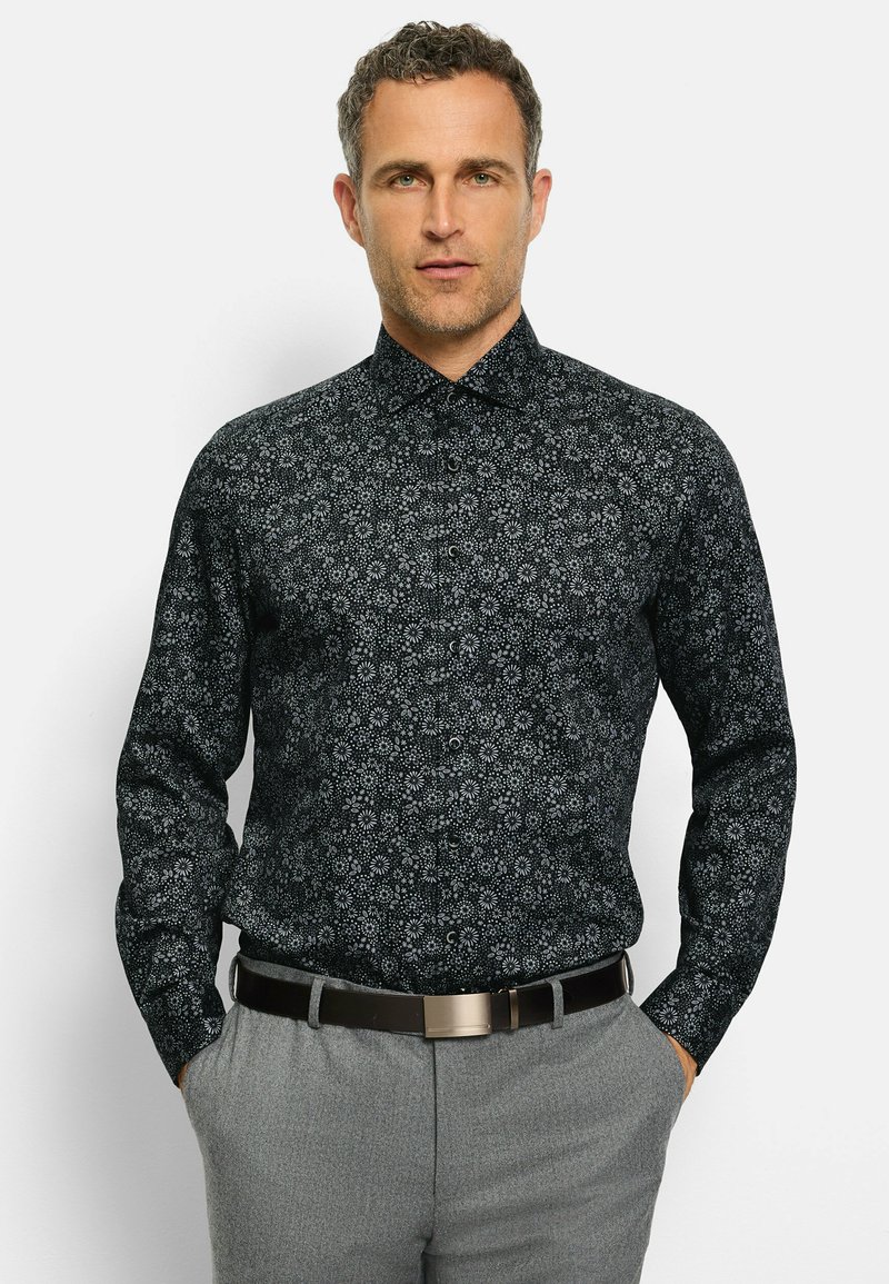 Schwarzes Langarmhemd mit weißem Blumenmuster, ergänzt durch einen klassischen Kragen und einen durchgehenden Knopfverschluss, getragen mit grauen Hosen und einem Gürtel.