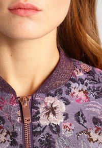 Fioletowa bomberka z kwiatowym wzorem, z metalicznym teksturowym kołnierzem i różowo-złotymi detalami zamka, z żywym wzorem różowych i białych kwiatów.