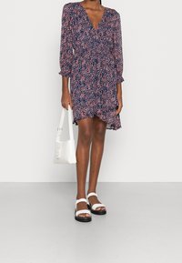 Marineblaues Blumenkleid mit pinken Akzenten, das einen tiefen V-Ausschnitt und Puffärmel hat. Kombiniert mit weißen Sandalen und einer kleinen weißen Umhängetasche.
