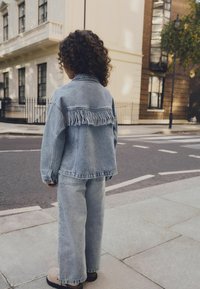 Jeansjacke mit Fransenverzierung auf dem Rücken, getragen über passenden weit geschnittenen Jeanshosen. Hellblau, lockere Passform, mit klassischem Kragen.