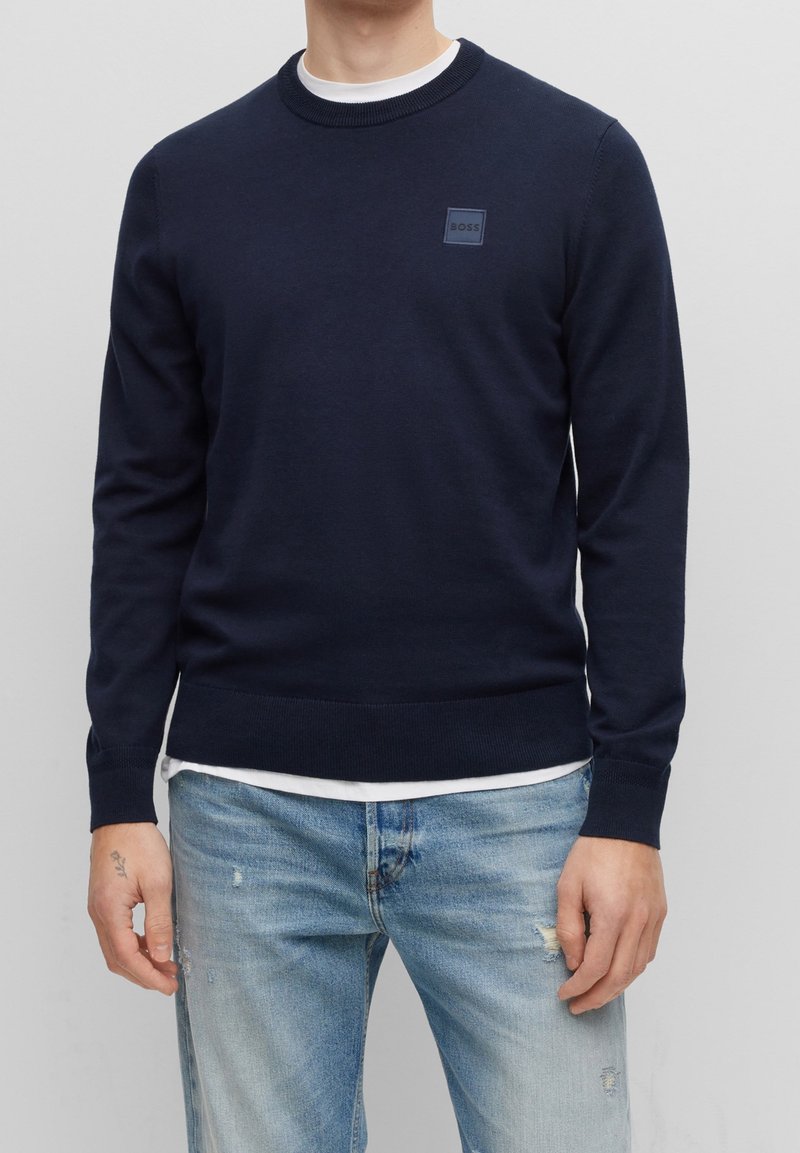 Navyblauwe gebreide trui met een ronde hals, geribde manchetten en zoom, en een klein vierkant logo op de borst, gedragen over een wit T-shirt.