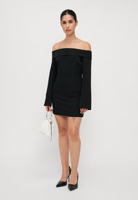 Svart off-the-shoulder kjole med lange ermer, tettsittende design, glatt stoff, kombinert med en hvit teksturert håndveske og svarte høye hæler.