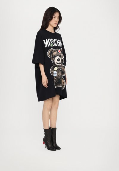 Μαύρο oversized φόρεμα t-shirt με γραφικό αρκουδάκι και παρασημειώσεις. Το μοντέλο φοράει μαύρες μπότες μέχρι τον αστράγαλο με κόκκινη λεπτομέρεια. Λείος υφασμάτινος τύπος.